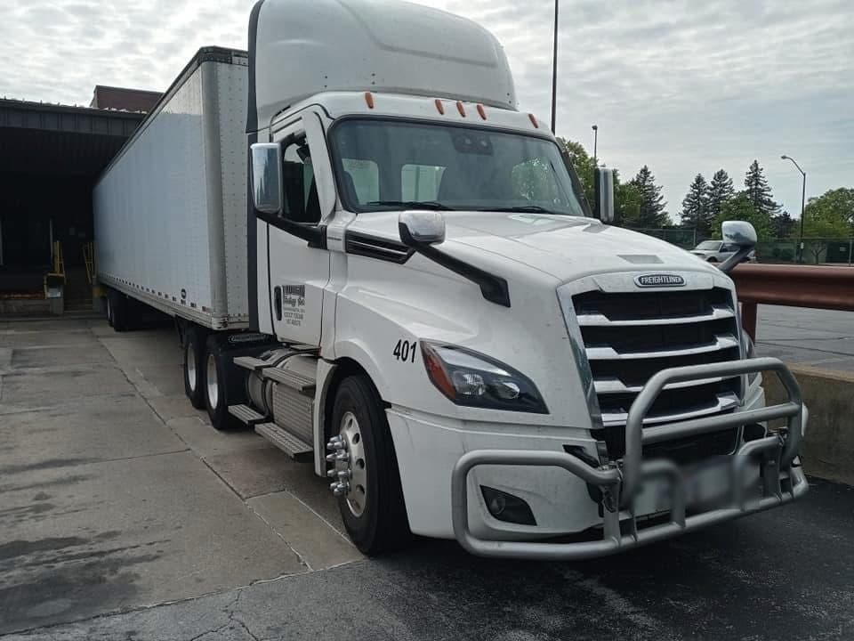 A white Freightliner Cascadia day cab with a heavy front bumper guard, coupled to a dry van at a dock.