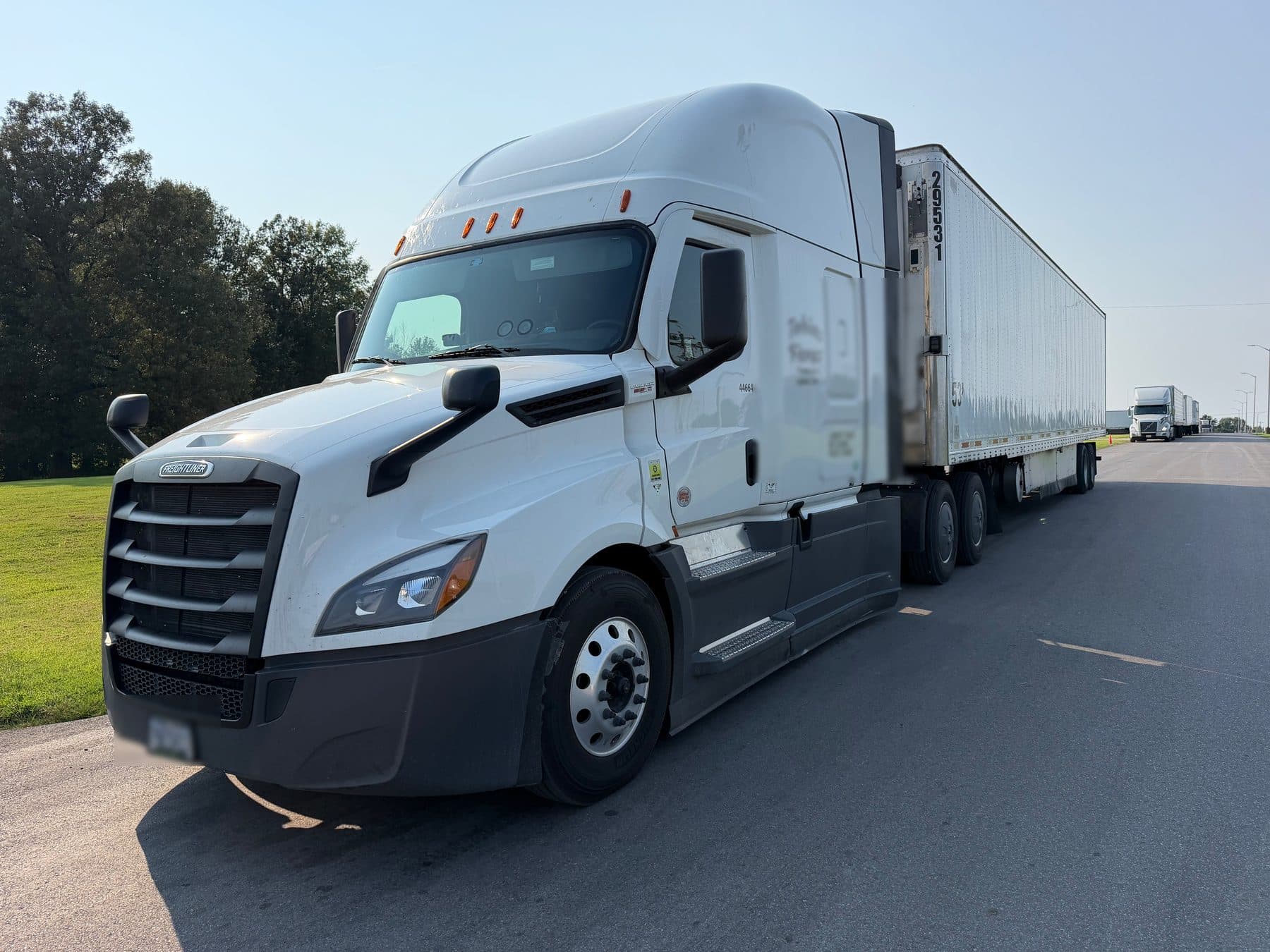 A white Freightliner Cascadia with a 53-foot refrigerated trailer, stopped on a grass shoulder in daylight.