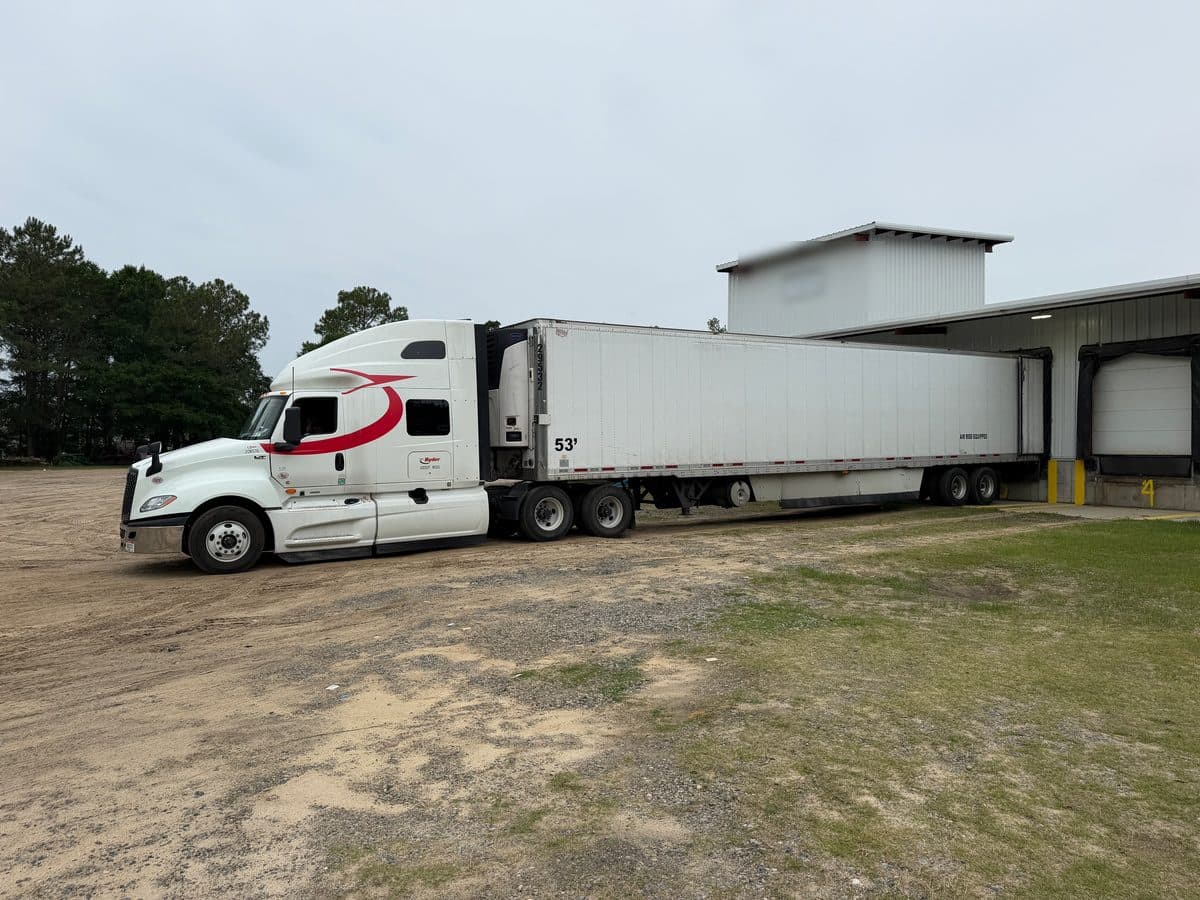 A white International sleeper and refrigerated trailer backed toward a warehouse dock on an overcast day.