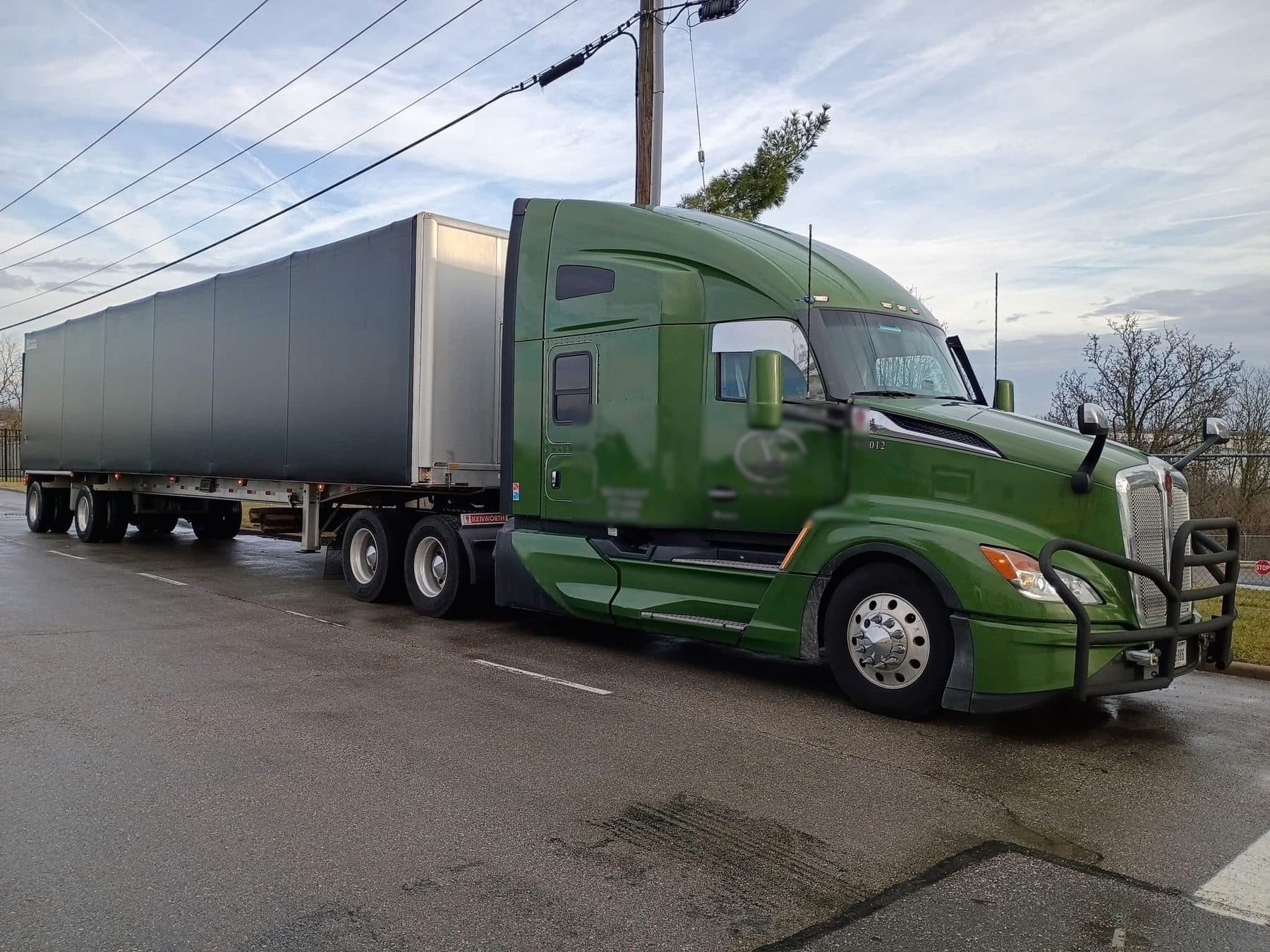A dark green Kenworth T680 pulling a Conestoga rolling-tarp trailer on wet pavement under a low sun.