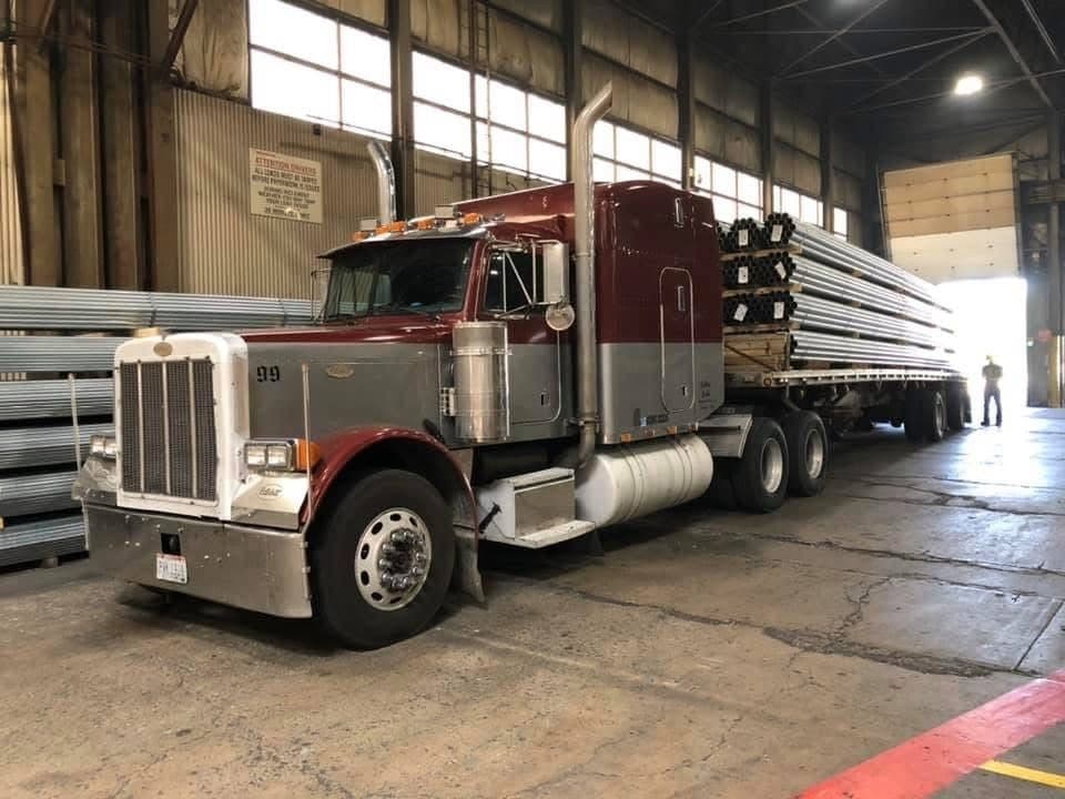 A maroon and silver Peterbilt 379 with a flatbed of bundled steel tubing, loading inside a mill.