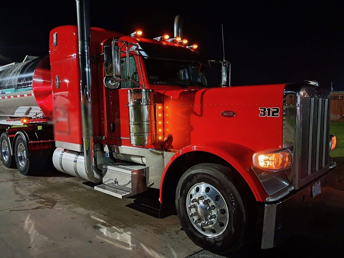 Close view of the front of a red Peterbilt 389 at night, cab lights lit, hitched to a stainless tanker.