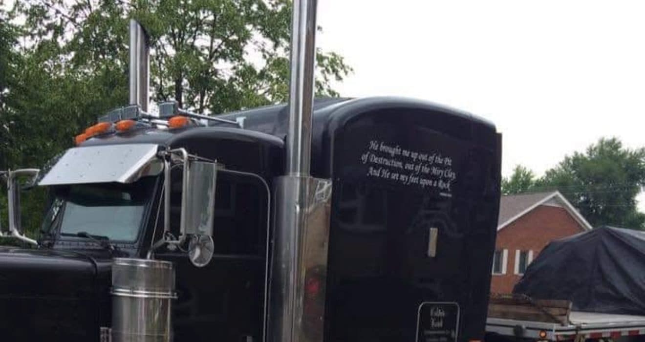 A black long-hood Peterbilt with chrome stacks and lettering across the sleeper, a tarped flatbed load behind it.