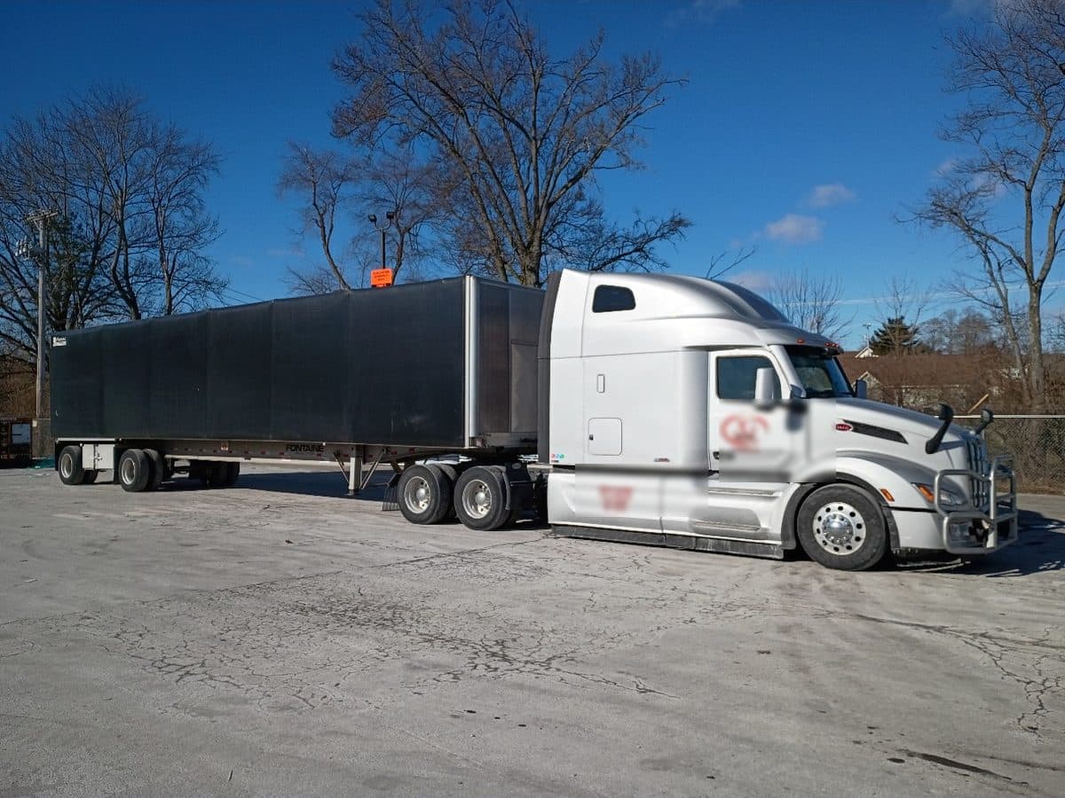 A white Peterbilt sleeper hitched to a Conestoga rolling-tarp flatbed on a clear winter day.